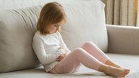 Little girl sitting on sofa and touching painful belly with arms at living room. Child suffering from stomach ache at home.