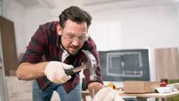 Hammering a nail into board. A carpenter wearing a red flannel shirt, jeans and cloth protective gloves nails the wooden boards